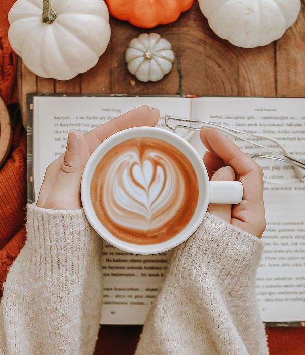 A lady’s hands wrapped around a coffee cup that has the foam shaped into a heart. She is holding her cup on top of a book with some white pumpkins next to it on a wooden table.